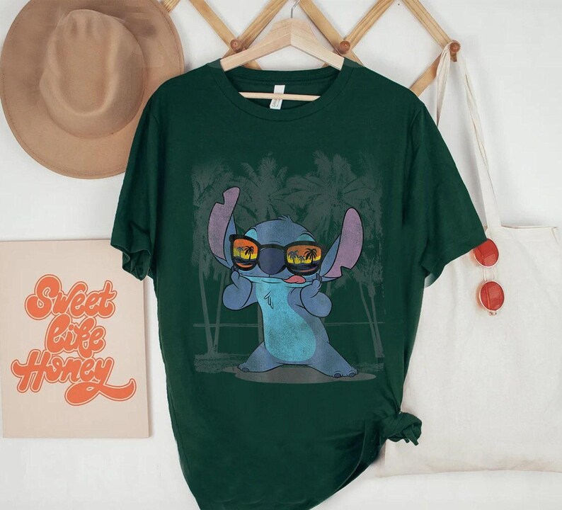Op de afbeelding: Een donkergroen T-shirt met korte mouwen en een afbeelding van een blauw alien-personage met een zonnebril waarin een zonsondergang en palmbomen weerspiegelen, tegen een achtergrond van vage palmbomsilhouetten.