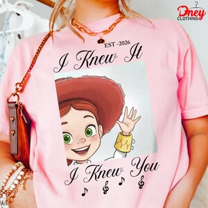Op de afbeelding: Een lichtroze t-shirt met een afbeelding van een stripfiguur met rood haar, een rode hoed en groene ogen. De tekst luidt "EST-2026", "I Knew It" en "I Knew You", met muzikale notensymbolen onder de illustratie van het personage.