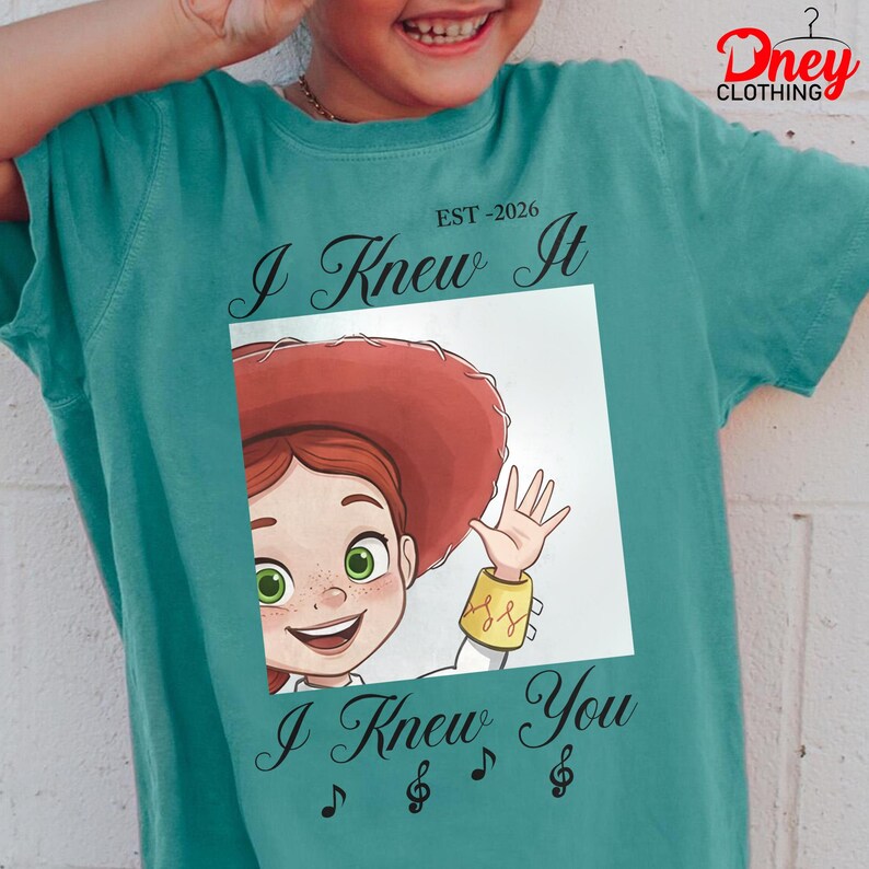 Op de afbeelding: Een blauwgroen t-shirt met korte mouwen en een afbeelding van een stripfiguur met rood haar en een rode hoed die zwaait. De tekst EST-2026, I Knew It en I Knew You, vergezeld door muzieknootsymbolen, is op de voorkant van het shirt gedrukt.