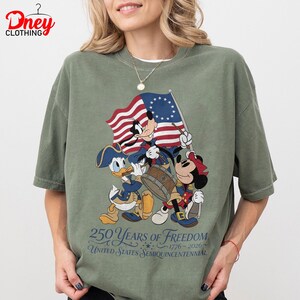 May include: Olive green t-shirt with Mickey Mouse, Donald Duck, and Minnie Mouse in patriotic outfits, holding a flag and drum. The shirt celebrates the United States Semiquincentennial, with the text "250 Years of Freedom 1776-2026."