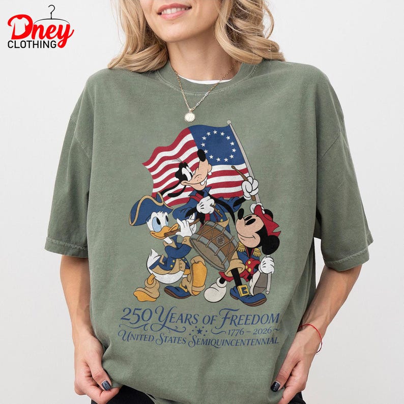 May include: Olive green t-shirt with Mickey Mouse, Donald Duck, and Minnie Mouse in patriotic outfits, holding a flag and drum. The shirt celebrates the United States Semiquincentennial, with the text "250 Years of Freedom 1776-2026."