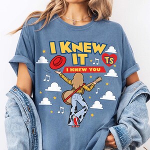 K&ouml;nnte beinhalten: Ein blaues T-Shirt mit einer Grafik einer Person mit langem Haar, die ein gelbes kariertes Hemd und eine Hose mit Kuhmuster tr&auml;gt und eine Gitarre h&auml;lt. Der Text lautet I KNEW IT und I KNEW YOU, erg&auml;nzt durch einen roten Hut, ein rotes Herz mit TS, Wolken und Musiknoten.