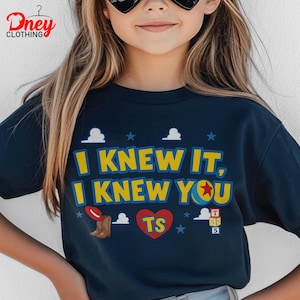 Puede incluir: Una camiseta azul marino oscuro con el texto amarillo I KNEW IT, I KNEW YOU. El dise&ntilde;o incluye un sombrero de vaquero rojo, una pelota amarilla con una estrella roja, un coraz&oacute;n rojo con las letras TS y tres bloques apilados con T, S y 5.