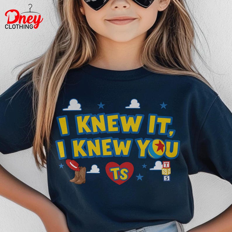 Puede incluir: Una camiseta azul marino oscuro con el texto amarillo I KNEW IT, I KNEW YOU. El dise&ntilde;o incluye un sombrero de vaquero rojo, una pelota amarilla con una estrella roja, un coraz&oacute;n rojo con las letras TS y tres bloques apilados con T, S y 5.