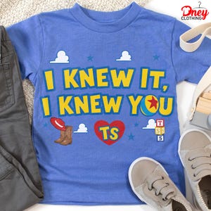 Puede incluir: Una camiseta azul de manga corta con el texto amarillo I KNEW IT, I KNEW YOU. El dise&ntilde;o incluye un sombrero de vaquero rojo, botas marrones, un coraz&oacute;n rojo con TS, una pelota con una estrella roja y tres bloques apilados con T, S y 5.
