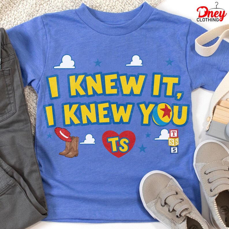 Puede incluir: Una camiseta azul de manga corta con el texto amarillo I KNEW IT, I KNEW YOU. El dise&ntilde;o incluye un sombrero de vaquero rojo, botas marrones, un coraz&oacute;n rojo con TS, una pelota con una estrella roja y tres bloques apilados con T, S y 5.