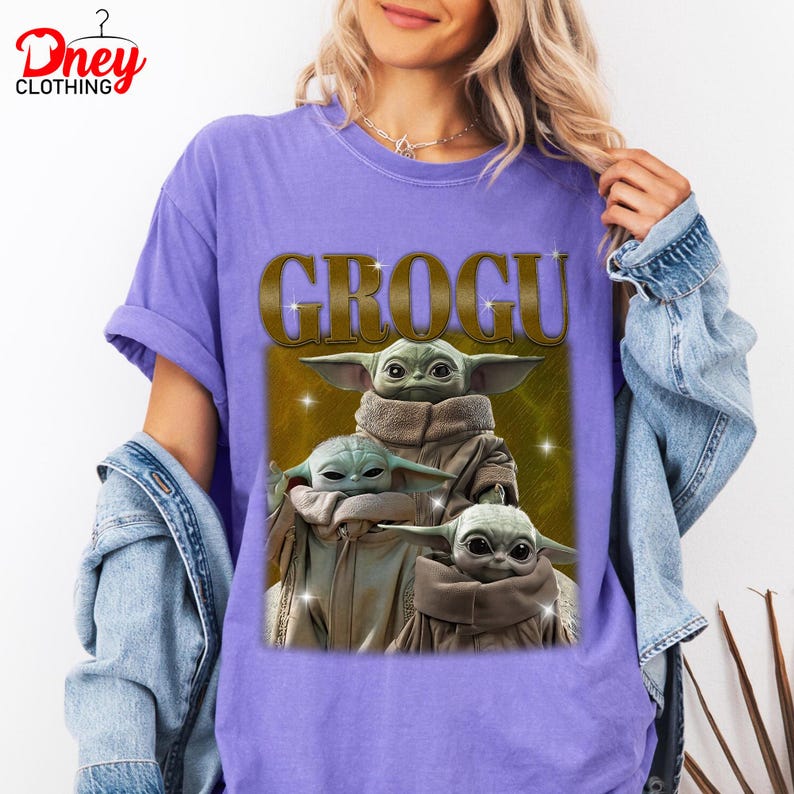 K&ouml;nnte beinhalten: Ein lila T-Shirt mit dem Wort "GROGU" in goldfarbenen Buchstaben und einer Grafik von drei Grogu-Charakteren. Die Grogu-Charaktere sind hellgr&uuml;n mit beigen Roben. Das T-Shirt hat kurze &Auml;rmel.