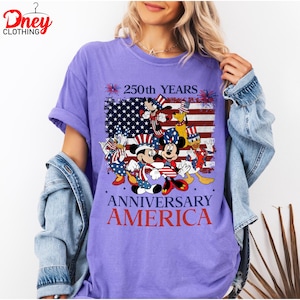 Puede incluir: Una camiseta morada con un gr&aacute;fico que presenta la bandera estadounidense y personajes de Disney con atuendos patri&oacute;ticos. La camiseta tiene el texto "250th YEARS ANNIVERSARY AMERICA". La camiseta es de cuello redondo y tiene mangas cortas.