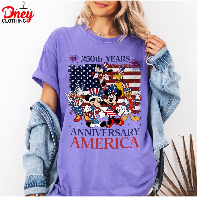 Puede incluir: Una camiseta morada con un gr&aacute;fico que presenta la bandera estadounidense y personajes de Disney con atuendos patri&oacute;ticos. La camiseta tiene el texto "250th YEARS ANNIVERSARY AMERICA". La camiseta es de cuello redondo y tiene mangas cortas.