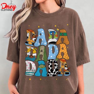 May include: Brown t-shirt with the word "DADA" in large, colorful, cartoon-style letters. The letters are filled with various patterns and character designs, including a cowboy boot and a cow print. Small yellow stars are scattered around the letters.