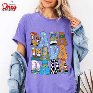 May include: A purple t-shirt with the word "DADA" in colorful, cartoon-style letters. The letters are designed with various patterns and character themes. The shirt has short sleeves and a relaxed fit.