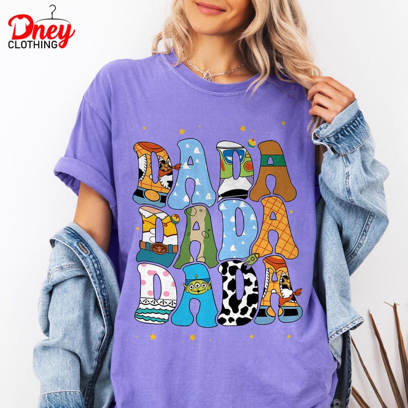 May include: A purple t-shirt with the word "DADA" in colorful, cartoon-style letters. The letters are designed with various patterns and character themes. The shirt has short sleeves and a relaxed fit.