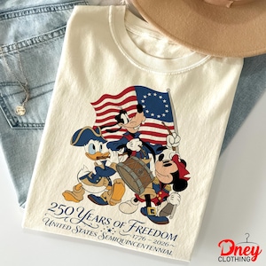 May include: Off-white t-shirt with Donald Duck, Goofy, and Mickey Mouse in patriotic outfits, holding a colonial American flag. The shirt celebrates 250 Years of Freedom, 1776-2026, United States Semiquincentennial.