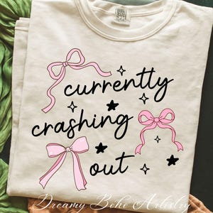 Puede incluir: Camiseta color crema con la frase "currently crashing out" en escritura negra, adornada con lazos rosas y estrellas negras. La frase "Dreamy Boho Artistry" está impresa en la parte inferior. Una camiseta informal y cómoda.