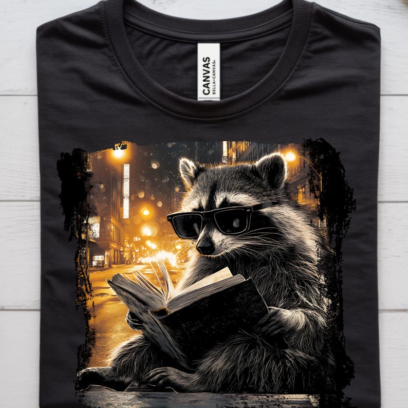 May include: Black t-shirt featuring a raccoon wearing sunglasses and reading a book. The raccoon is set against a blurred background of a city street at night, with warm yellow streetlights. The shirt has a crew neck and the brand "Canvas" is visible.