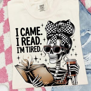 Może przedstawiać: Kremowa koszulka z napisem "I CAME. I READ. I'M TIRED." Szkielet w okularach przeciwsłonecznych, w szachową kokardą i filiżanką kawy czyta książkę z płomieniem.