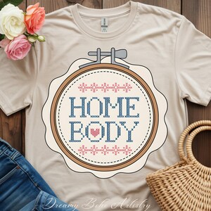 May include: Cream-coloured t-shirt with a cross-stitch design in a wooden embroidery hoop. The design spells "HOME BODY" in blue, with a pink heart. The top is a Gildan brand. The words "Dreamy Boho Artistry" are at the bottom.