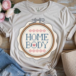 May include: A beige t-shirt with a cross-stitch design that reads "HOME BODY" in blue, with a red heart and pink and blue decorative elements. The design is framed in a round embroidery hoop. The shirt is folded with a pair of blue jeans.