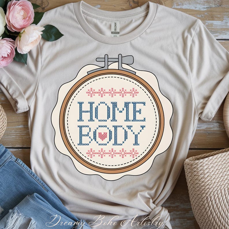 May include: A beige t-shirt with a cross-stitch design that reads "HOME BODY" in blue, with a red heart and pink and blue decorative elements. The design is framed in a round embroidery hoop. The shirt is folded with a pair of blue jeans.