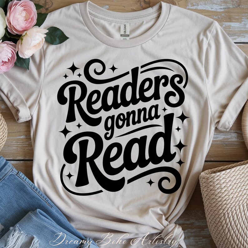 K&ouml;nnte beinhalten: Beiges T-Shirt mit dem schwarzen Schriftzug "Readers gonna Read" in einem Retro-Stil mit Sternen. Das Shirt ist gefaltet, mit blauen Jeans und Blumen im Hintergrund.