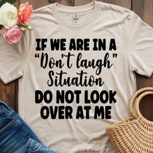 May include: A beige t-shirt with the text "IF WE ARE IN A 'Don't laugh' Situation DO NOT LOOK OVER AT ME" in black. The shirt is displayed with a pair of jeans, flowers, and a woven basket.