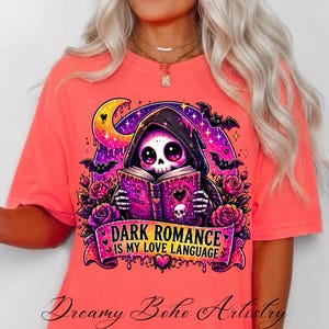 May include: Coral-colored t-shirt featuring a graphic of a skeleton in a hooded cloak reading a book. The design includes bats, roses, and the text "DARK ROMANCE IS MY LOVE LANGUAGE". The shirt is from Dreamy Boho Artistry.