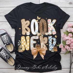 May include: Black t-shirt with the words "BOOK NERD" in a decorative font. The letters are filled with various patterns, including floral, leopard print, and script. A large bow and butterflies accent the design. The text "Dreamy Boho Artistry" is at the bottom.