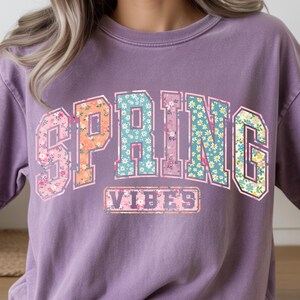 May include: A purple sweatshirt with the words "SPRING VIBES" in a floral design. The word "SPRING" is in large, block letters with a floral pattern in pink, orange, and blue. "VIBES" is in a smaller, rectangular box.