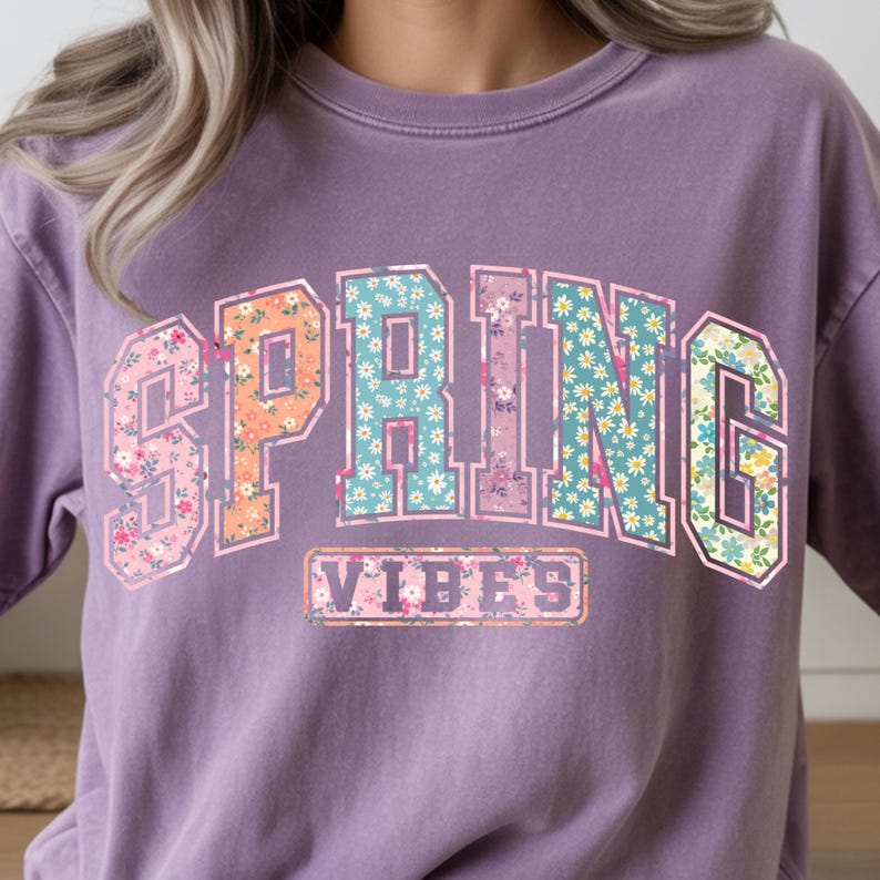 May include: A purple sweatshirt with the words "SPRING VIBES" in a floral design. The word "SPRING" is in large, block letters with a floral pattern in pink, orange, and blue. "VIBES" is in a smaller, rectangular box.