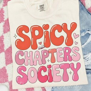 May include: Cream-coloured t-shirt featuring the text "SPICY CHAPTERS SOCIETY" in a retro font. The words are in shades of red and pink, with heart and star accents. The shirt is folded on denim.