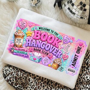 May include: White sweatshirt with a colorful graphic design. The design features the text "WELCOME TO THE BOOK HANGOVER BOOK CLUB" in pink and purple, along with illustrations of coffee, books, and hearts. The words "ADMIT ONE" are also visible.