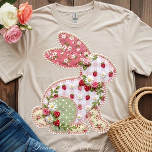May include: A beige t-shirt featuring a decorative bunny design. The bunny is made of various patterned fabrics, including pink floral, gingham, and green polka dots, with embroidered details, strawberries, and daisies. The text "Dreamy Boho Artistry" is at the bottom.