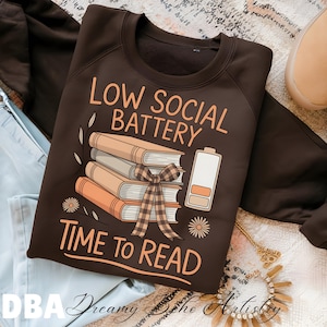 Puede incluir: Una sudadera marrón con el texto "LOW SOCIAL BATTERY TIME TO READ" en naranja. El diseño incluye una pila de libros atados con una cinta a cuadros y un icono de batería baja. La sudadera es de estilo casual.