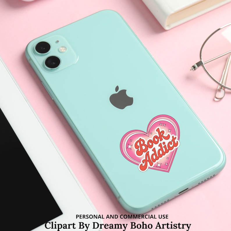 Op de afbeelding: Een lichtblauwe smartphone met een roze en rode hartvormige sticker met de tekst "Book Addict". De telefoon heeft een zwart Apple-logo en een camera met dubbele lens. De sticker heeft een retro-ontwerp.
