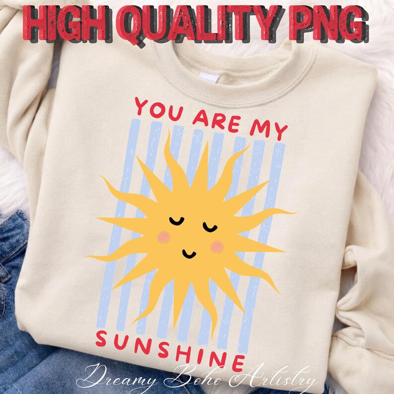 May include: Cream-colored sweatshirt with a smiling yellow sun graphic and the text "YOU ARE MY SUNSHINE" in red. The sun is centered on a light blue striped background. The words "Dreamy Boho Artistry" are at the bottom.