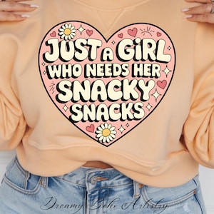May include: A peach-colored sweatshirt with a pink heart graphic. The heart graphic features the text "Just a girl who needs her snacky snacks" in a retro font with white outlines. The graphic also includes small hearts, stars, and flowers.