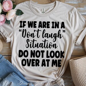 May include: A beige t-shirt with the text "IF WE ARE IN A 'Don't laugh' Situation DO NOT LOOK OVER AT ME" in black. The shirt is styled with a pair of blue jeans and a woven basket. Floral accents are visible on the left side.