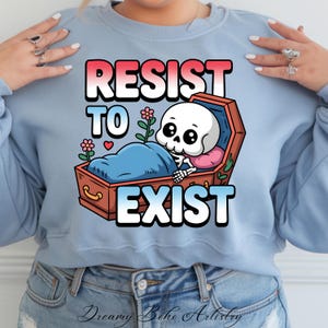 以下が含まれることがあります： 薄い青色のスウェットシャツに、花と「Resist to Exist」という文字が書かれた棺桶の中の骸骨のグラフィックがプリントされています。