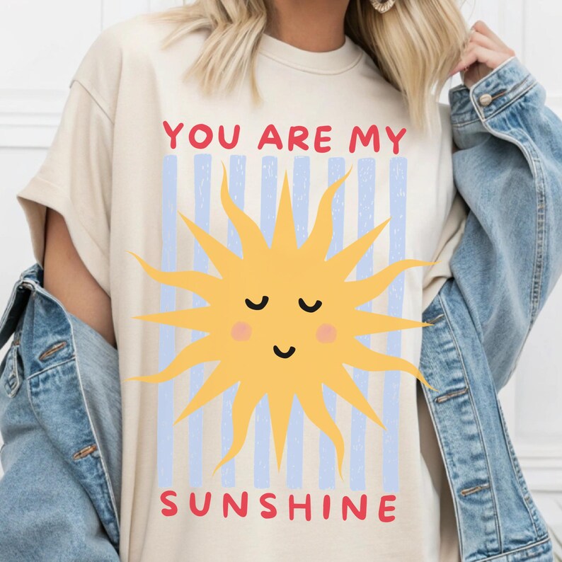 May include: Cream-colored t-shirt with a smiling yellow sun graphic and the text "YOU ARE MY SUNSHINE" in red. The design includes blue vertical stripes. A casual, comfortable top.