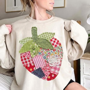Può includere: Felpa color crema con un grande design a patchwork a forma di fragola. La fragola è composta da vari modelli di tessuto, tra cui quadretti, fiori e toppe a righe in rosso, rosa, verde e blu. Le foglie e il gambo sono verdi.