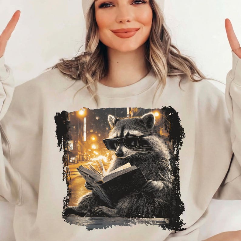 May include: Beige sweatshirt featuring a graphic of a raccoon wearing sunglasses and reading a book. The raccoon is set against a blurred city street scene with streetlights. The sweatshirt is a neutral color.
