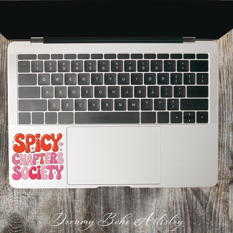 May include: A laptop with a sticker reading "SPICY CHAPTERS SOCIETY" in red and pink bubble letters. The laptop is open on a wooden surface. The keyboard is visible.