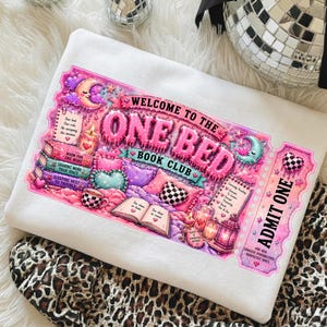 May include: White sweatshirt featuring a colorful graphic of a ticket that reads "Welcome to the One Bed Book Club." The design includes a bed, books, hearts, and the text "Admit One."