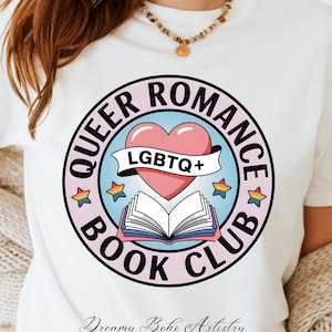 Op de afbeelding: Wit T-shirt met een cirkelvormig ontwerp met de tekst "QUEER ROMANCE BOOK CLUB". Het ontwerp bevat een roze hart, een open boek en de tekst "LGBTQ+". Kleurrijke sterren en een lichtblauwe achtergrond maken het ontwerp compleet.