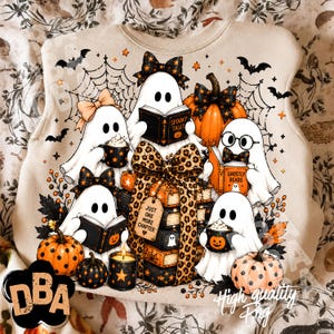 May include: A beige sweatshirt featuring a graphic of five white ghosts, pumpkins, and a stack of books tied with a leopard print bow. The books include text reading Spooky Tales, Ghostly Reads, and a tag that says Just One More Chapter.