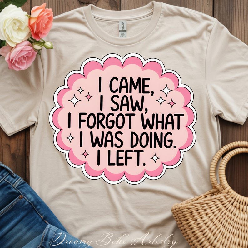 May include: A beige t-shirt with the text "I CAME, I SAW, I FORGOT WHAT I WAS DOING. I LEFT." in a pink and white design. The design is surrounded by a pink scalloped border and star accents. The shirt is displayed with a floral arrangement and a woven basket.