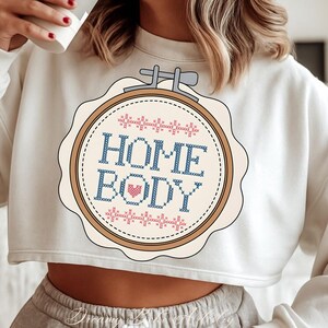 May include: Cream-coloured cropped sweatshirt with a cross-stitch design. The design features the words "HOME BODY" in blue, a pink heart, and pink and blue decorative stitching. The design is in a round, scalloped frame.