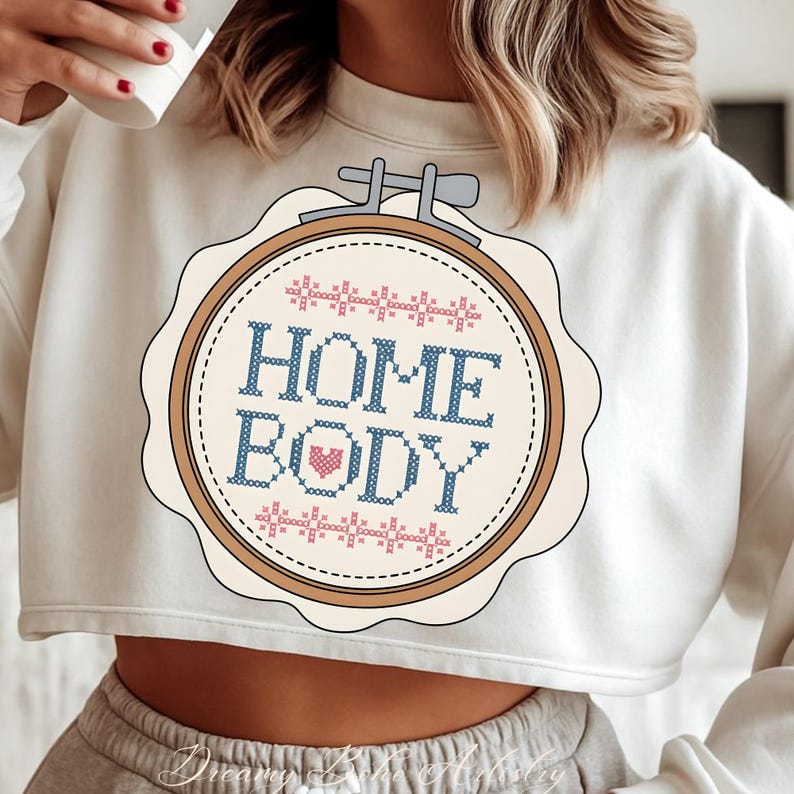 May include: Cream-coloured cropped sweatshirt with a cross-stitch design. The design features the words "HOME BODY" in blue, a pink heart, and pink and blue decorative stitching. The design is in a round, scalloped frame.