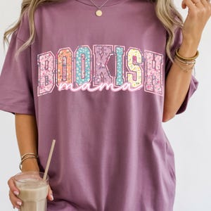 May include: A mauve-colored t-shirt with the text "BOOKISH mama" in a colorful floral design. The letters are outlined in a light pink and filled with various floral patterns. The shirt has short sleeves.