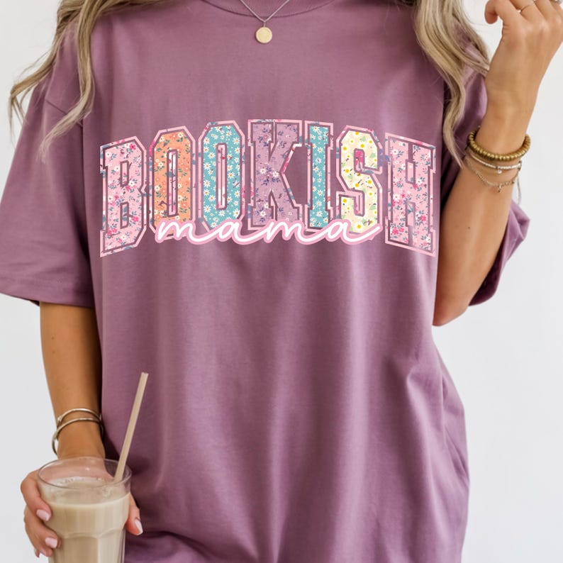May include: A mauve-colored t-shirt with the text "BOOKISH mama" in a colorful floral design. The letters are outlined in a light pink and filled with various floral patterns. The shirt has short sleeves.